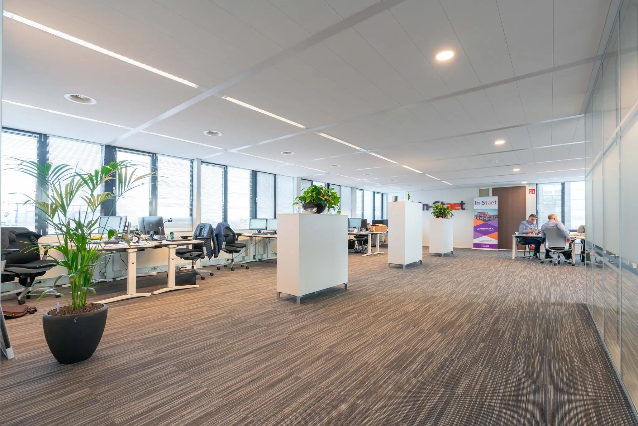 Modern kantoorinterieur aan de De Bruyn Kopsstraat met werkplekken, planten en medewerkers in overleg.